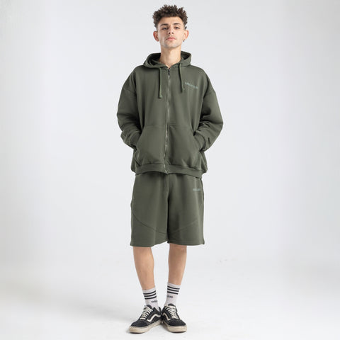 Model wearing the BROWNTOAST Core Long Shorts from the Core Drop in olive green — relaxed unisex cotton-lycra shorts with front piping detail, side pockets, and an adjustable drawstring waist. Knee-length design balancing bermuda and short fit, made in Egypt for everyday comfort and style.