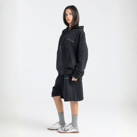 Model wearing the BROWNTOAST Core Long Shorts from the Core Drop in black — relaxed unisex cotton-lycra shorts with front piping detail, side pockets, and an adjustable drawstring waist. Knee-length design balancing bermuda and short fit, made in Egypt for everyday comfort and style.