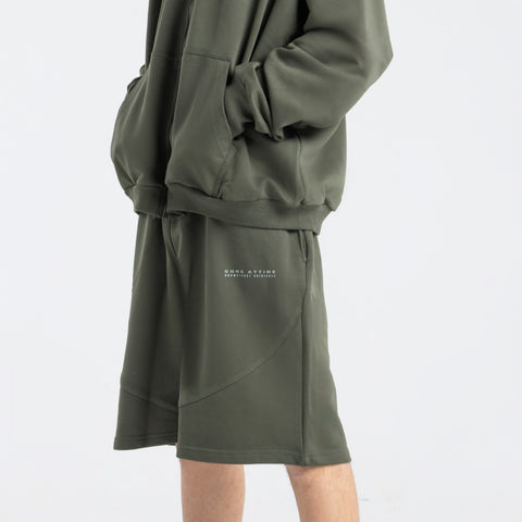 Model wearing the BROWNTOAST Core Long Shorts from the Core Drop in olive green — relaxed unisex cotton-lycra shorts with front piping detail, side pockets, and an adjustable drawstring waist. Knee-length design balancing bermuda and short fit, made in Egypt for everyday comfort and style.