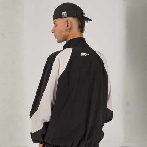 Model wearing the BROWNTOAST Compton Tracksuit Jacket — unisex oversized polyester track jacket with a dual-function zipper and BTO rubber and embroidered logos. Inspired by old-school Compton style and 80s hip-hop culture. Available in black and white.